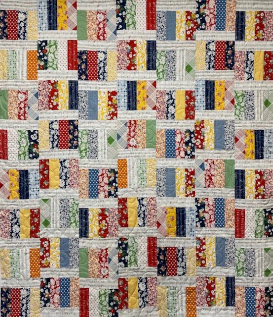 A colorful quilt featuring a patchwork design with various patterns and fabrics, arranged in blocks of different colors including red, blue, yellow, green, and floral prints.
