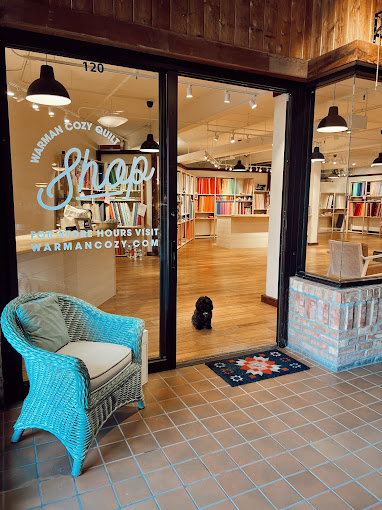A cozy quilt shop entrance featuring a glass door with the words 'Warman Cozy Quilt Shop' and a decorative blue chair on the porch. A small black dog sits in front of the entrance, adding charm to the scene.