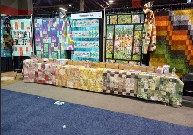 A booth display at the H&H Americas Show featuring colorful quilts and fabric designs by Villa Rose Designs, with a table showcasing various quilting products.