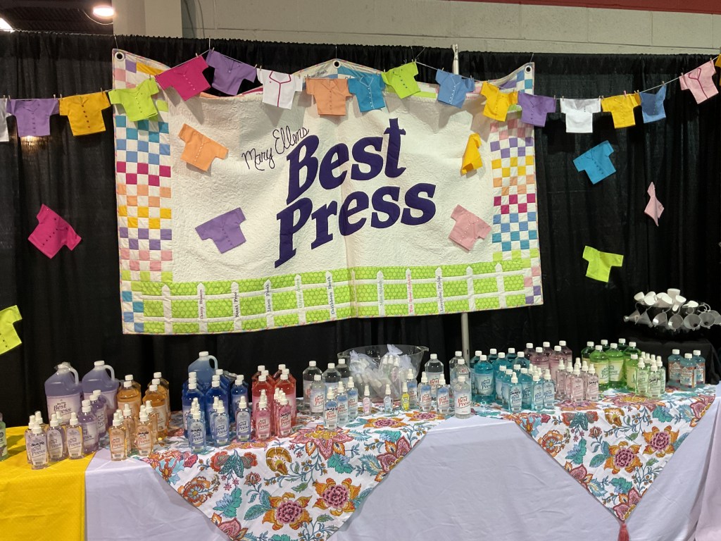 A booth display for Mary Ellen's Best Press at a quilting show, featuring colorful quilted decorations, various sizes of spray bottles, and a floral tablecloth.