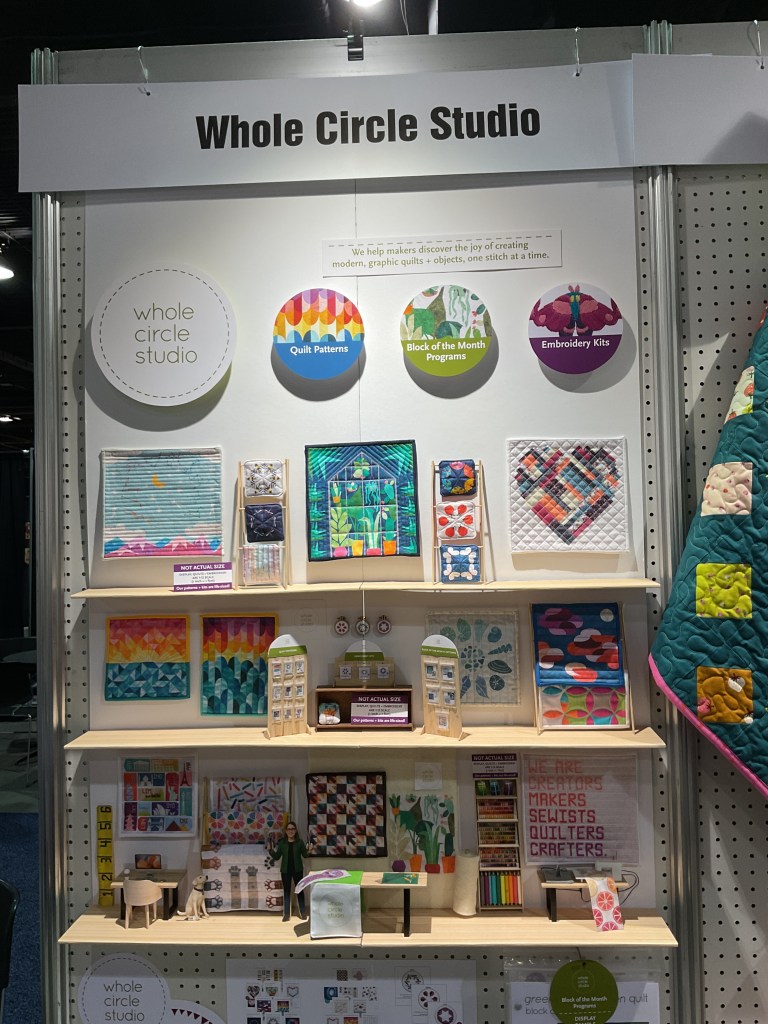 Display of Whole Circle Studio showcasing colorful quilt patterns, embroidery kits, and a miniature quilt shop setup.