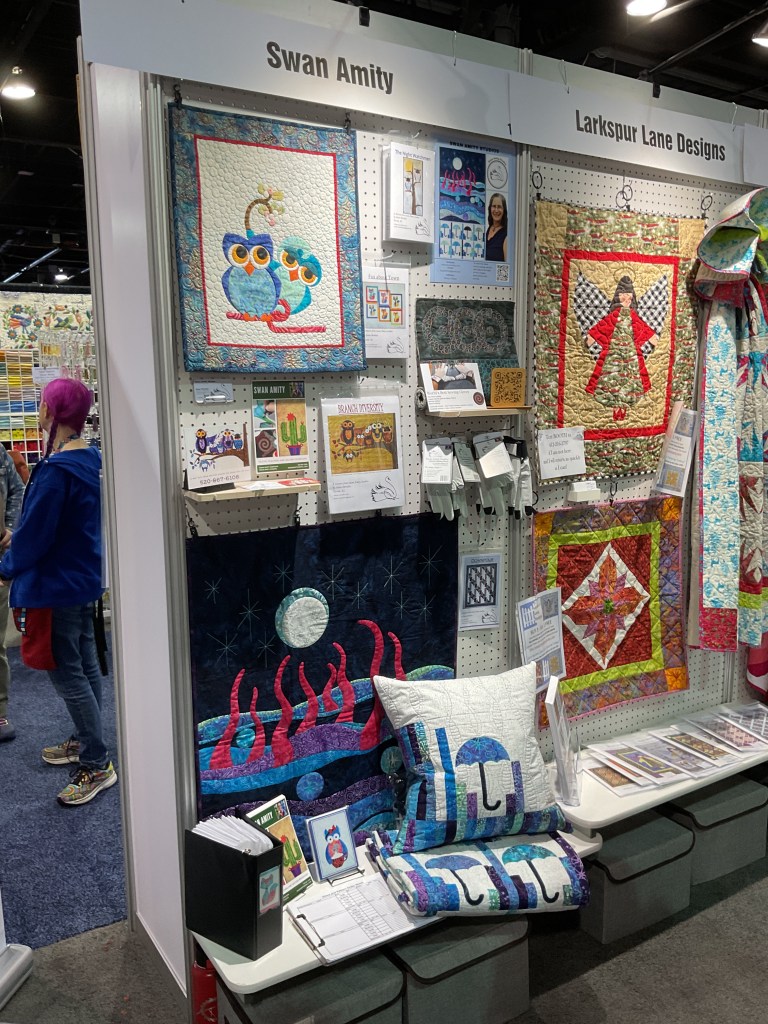 A colorful quilting booth featuring designs by Swan Amity and Larkspur Lane Designs, showcasing various quilted items, including wall hangings and pillows, with a vibrant display of fabrics and patterns.