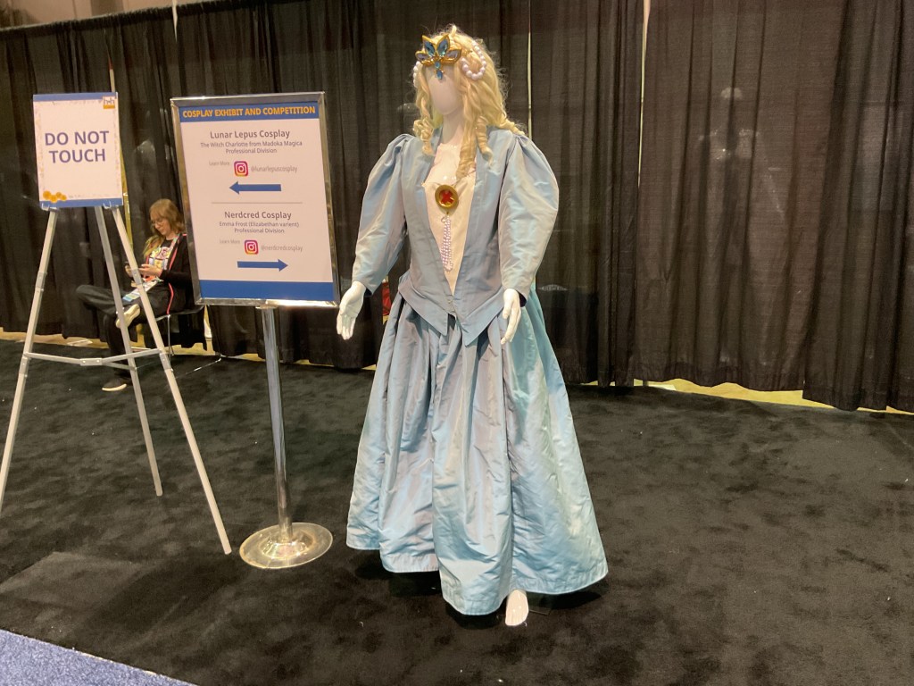 A mannequin dressed in an elaborate light blue gown representing a cosplay character, displayed next to a sign indicating the cosplay exhibit and competition categories.
