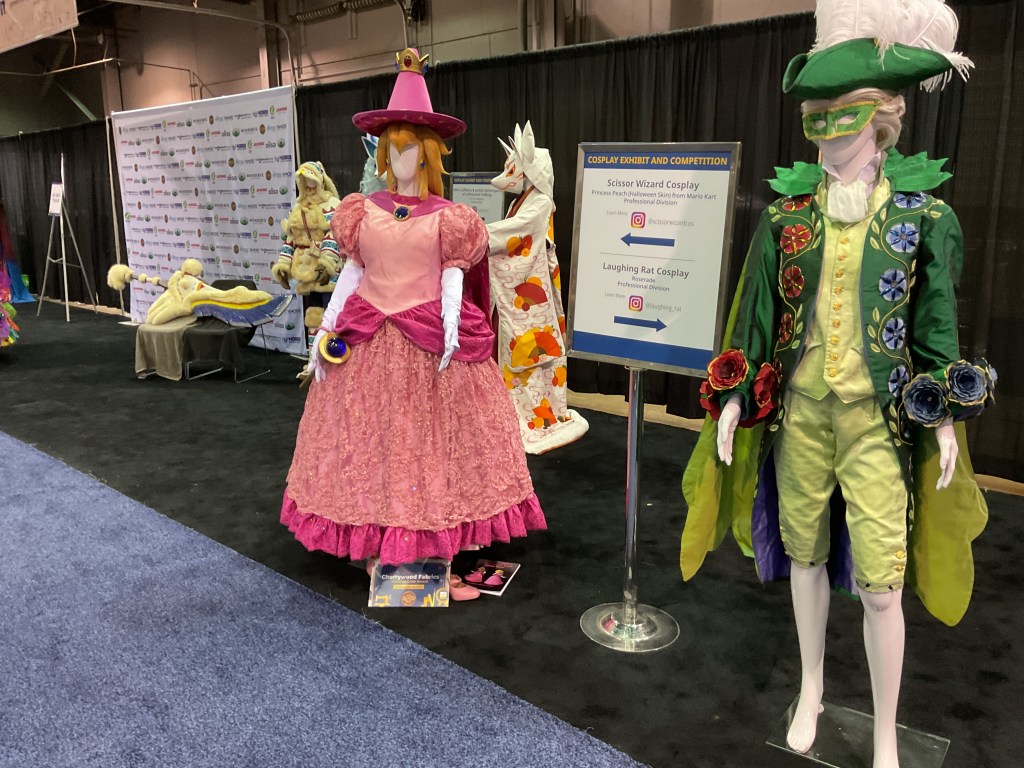 Cosplay exhibit featuring two mannequins dressed in vibrant costumes. The left mannequin is wearing a pink dress with puffed sleeves and a crown, while the right mannequin is in a colorful suit with floral accents and a mask. In the background, a sign indicates 'Scissor Wizard Cosplay' and 'Laughing Rat Cosplay'.