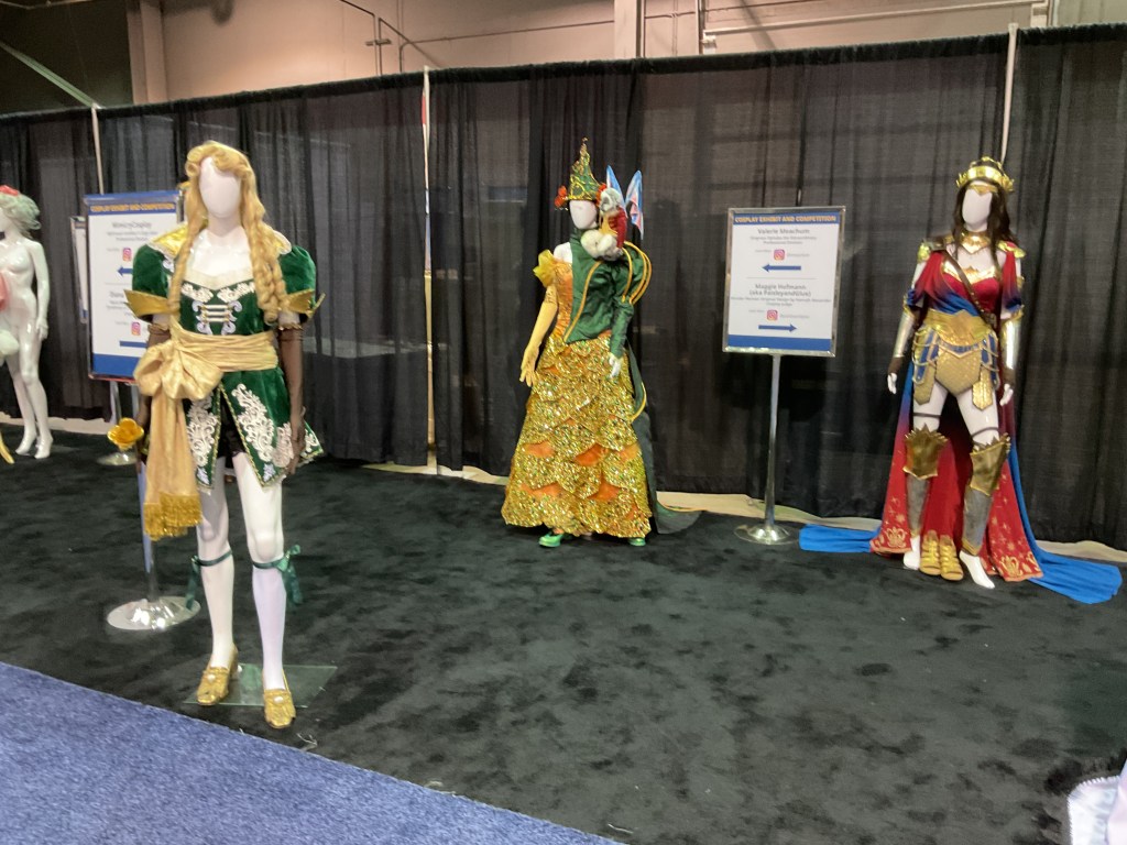 Three cosplay costumes displayed on mannequins at a convention, featuring vibrant colors and intricate designs.