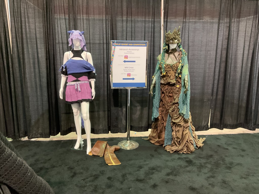 Two cosplay outfits on display at a competition, one featuring a colorful top and shorts, and the other resembling a nature-inspired costume with green and brown tones.