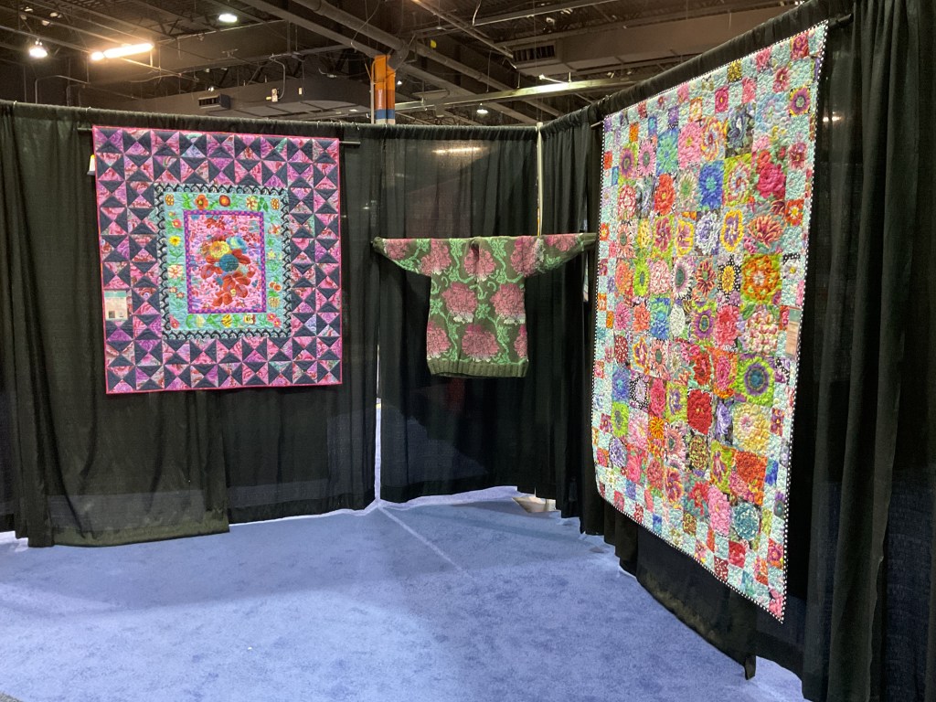A vibrant display of quilts featuring colorful patterns, including a wall quilt with floral designs and a garment hanging among the fabric backdrop at a quilting show.