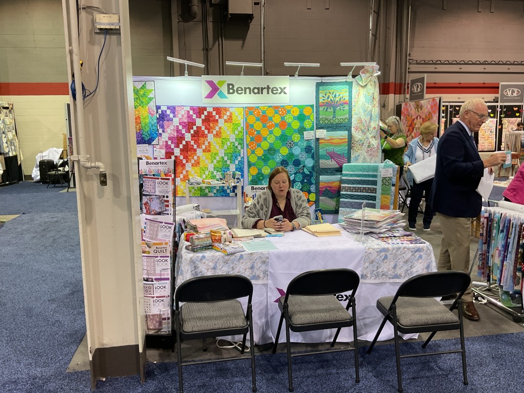A booth at the H&H Americas Show featuring colorful quilt displays, with a woman sitting at the table and attendees browsing nearby.