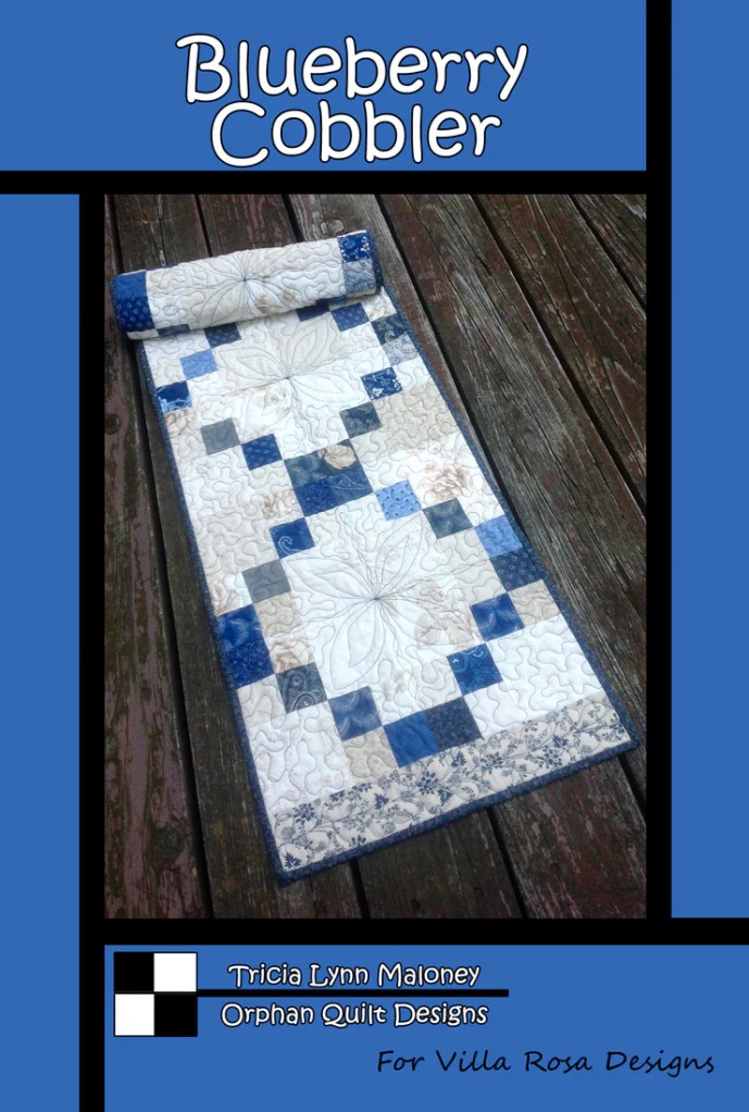 A rolled quilt titled 'Blueberry Cobbler' featuring a patchwork design in blue and cream colors, displayed on a wooden surface.