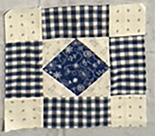 A patchwork quilt block featuring a central diamond shape in navy blue surrounded by light-colored fabric with small patterns and a checked design.