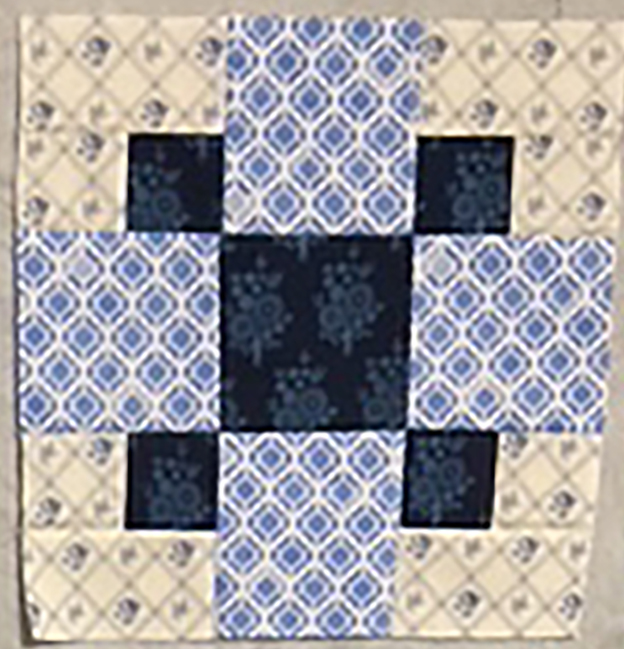 A square quilt block featuring a mix of blue and cream fabric patterns, arranged in a cross design with floral and geometric prints.