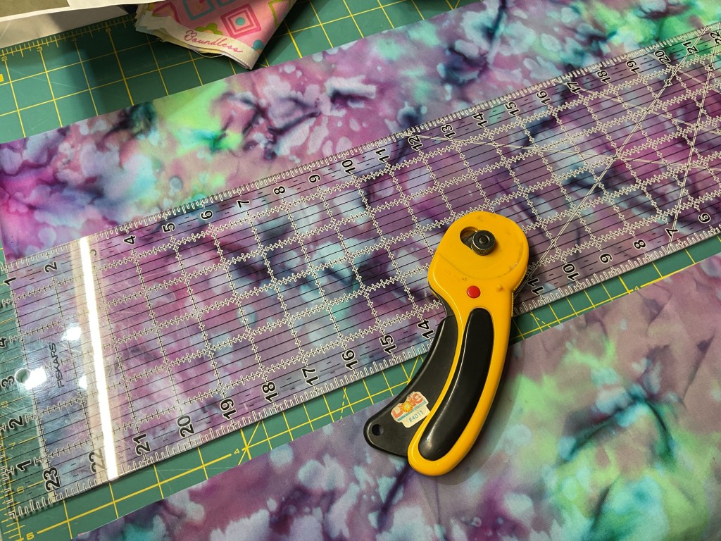 Close-up of fabric and quilting tools on a cutting mat, featuring a rotary cutter and a clear ruler positioned on colorful batik fabric.
