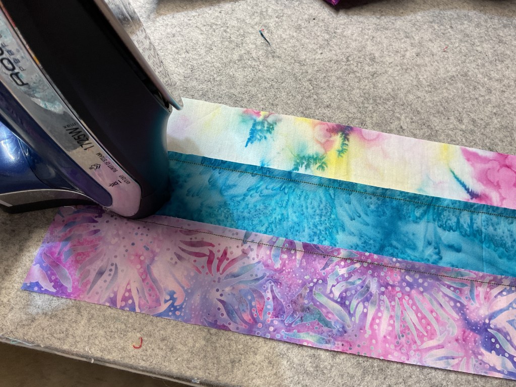 An iron placed on top of several colorful fabric strips, with a light blue, turquoise, and pink batik design visible.