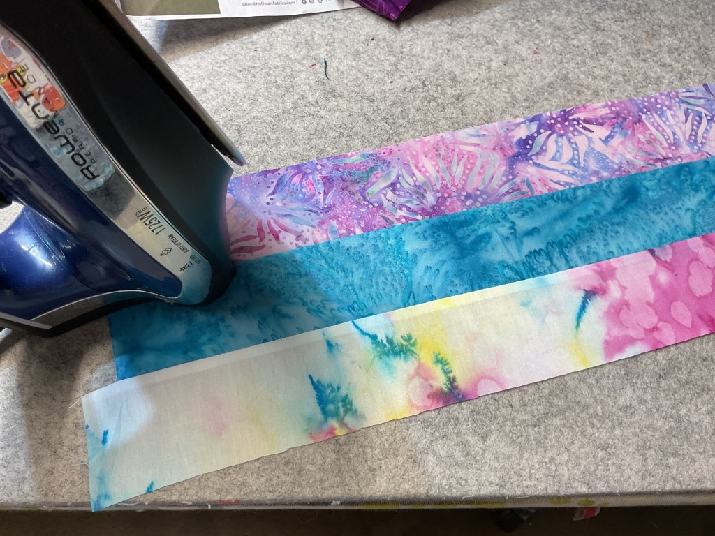 An iron rests on top of several colorful batik fabric strips arranged in a row on a cutting mat.