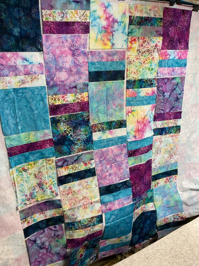 A vibrant quilt top featuring an arrangement of colorful batik fabrics in various shades of purple, blue, and green, organized in a patchwork pattern.