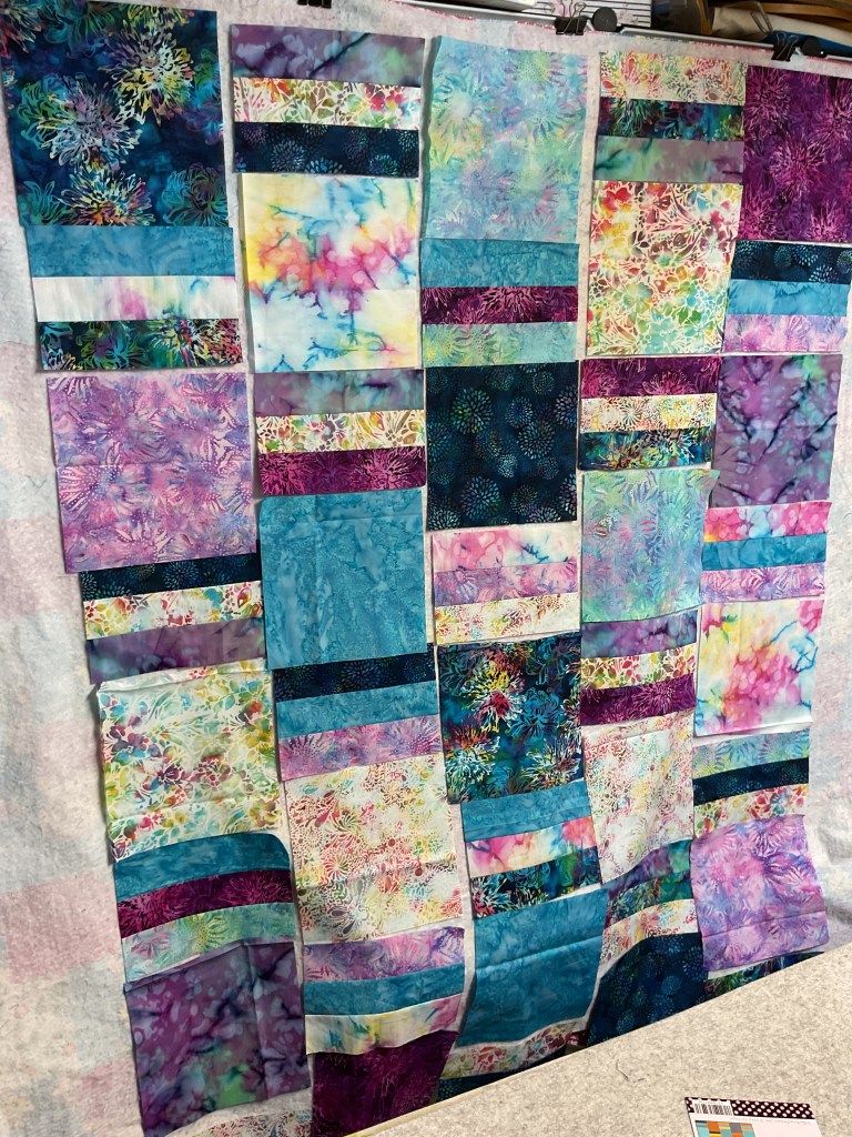 A colorful layout of batik fabric pieces arranged to form a quilt top, featuring a mix of blue, purple, and pastel shades, displayed on a design wall.