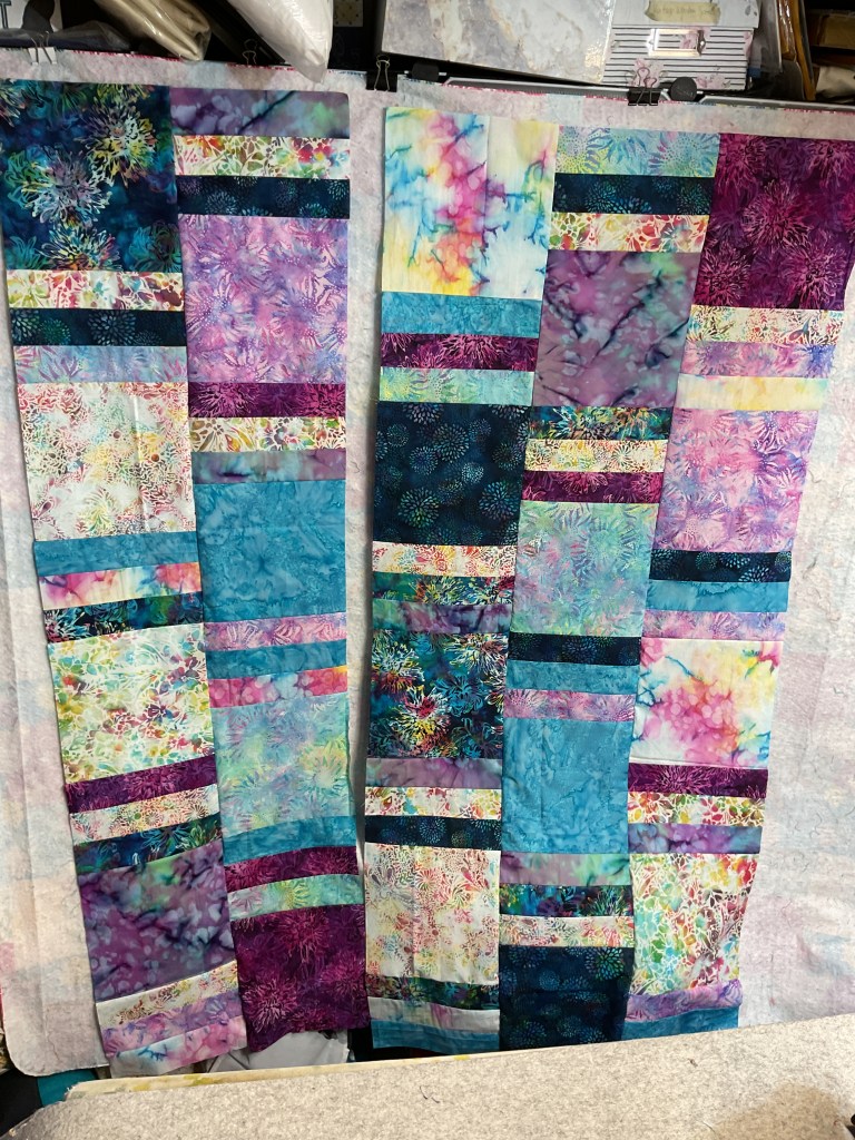 Two sections of a colorful, batik quilt featuring various shades of blue, purple, and colorful patterns, displayed on a design wall.