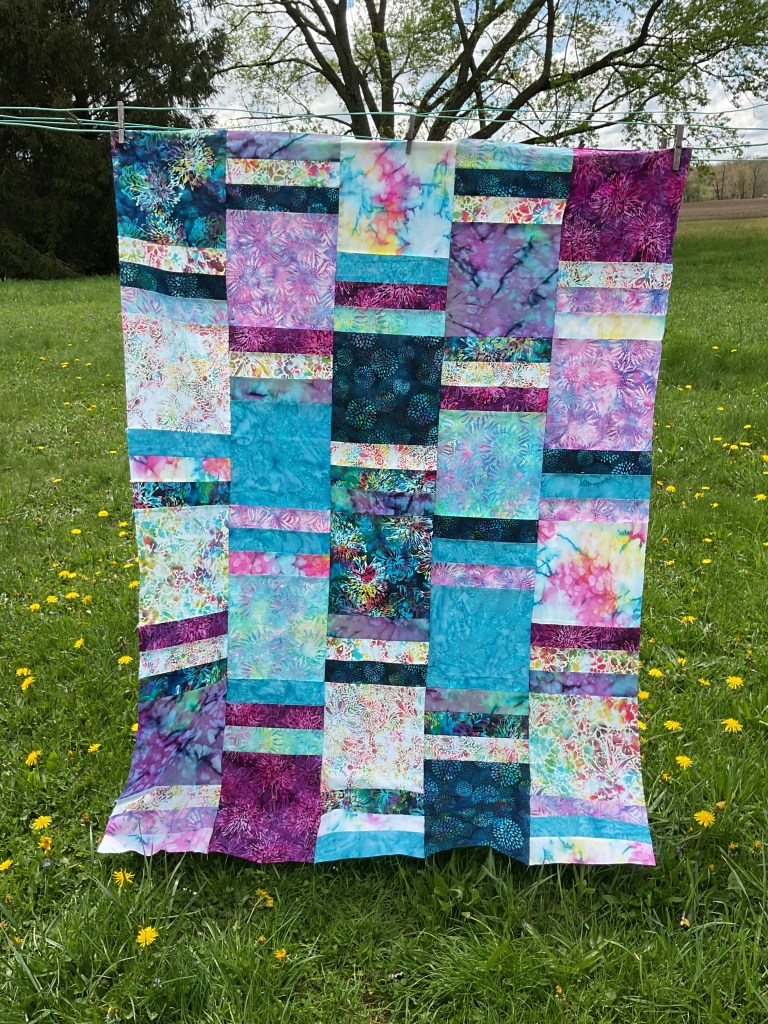 A colorful quilt made with batik fabrics, hanging on a clothesline outdoors against a backdrop of green grass and trees.