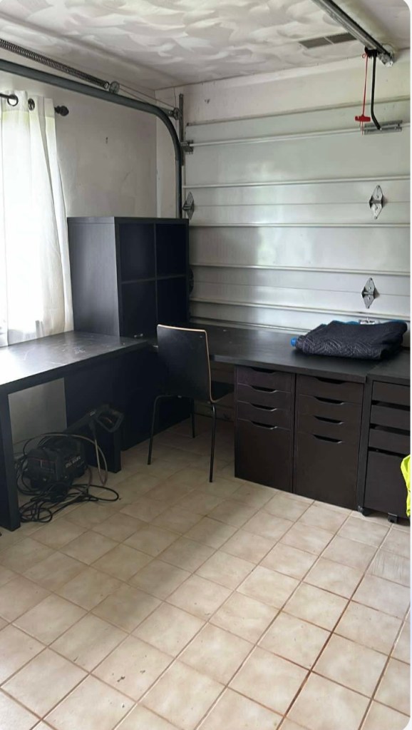 A garage space showing a dark brown office desk setup, including a desk, shelving unit, and a chair, with tiles on the floor and natural light coming through a window.