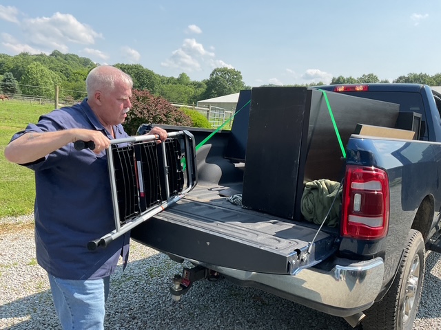 A man loading furniture into the back of a blue truck, with a sunny sky and greenery in the background.