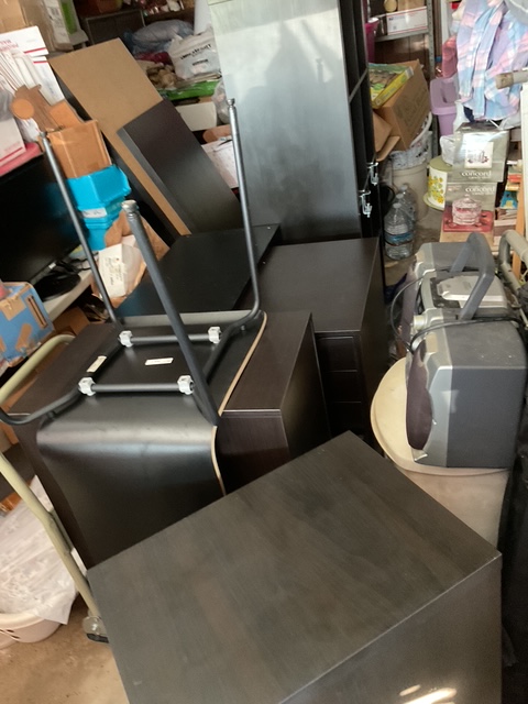 A cluttered workspace filled with dark brown Ikea office furniture pieces, including desktops, drawer units, and a chair, ready to be assembled in a studio.