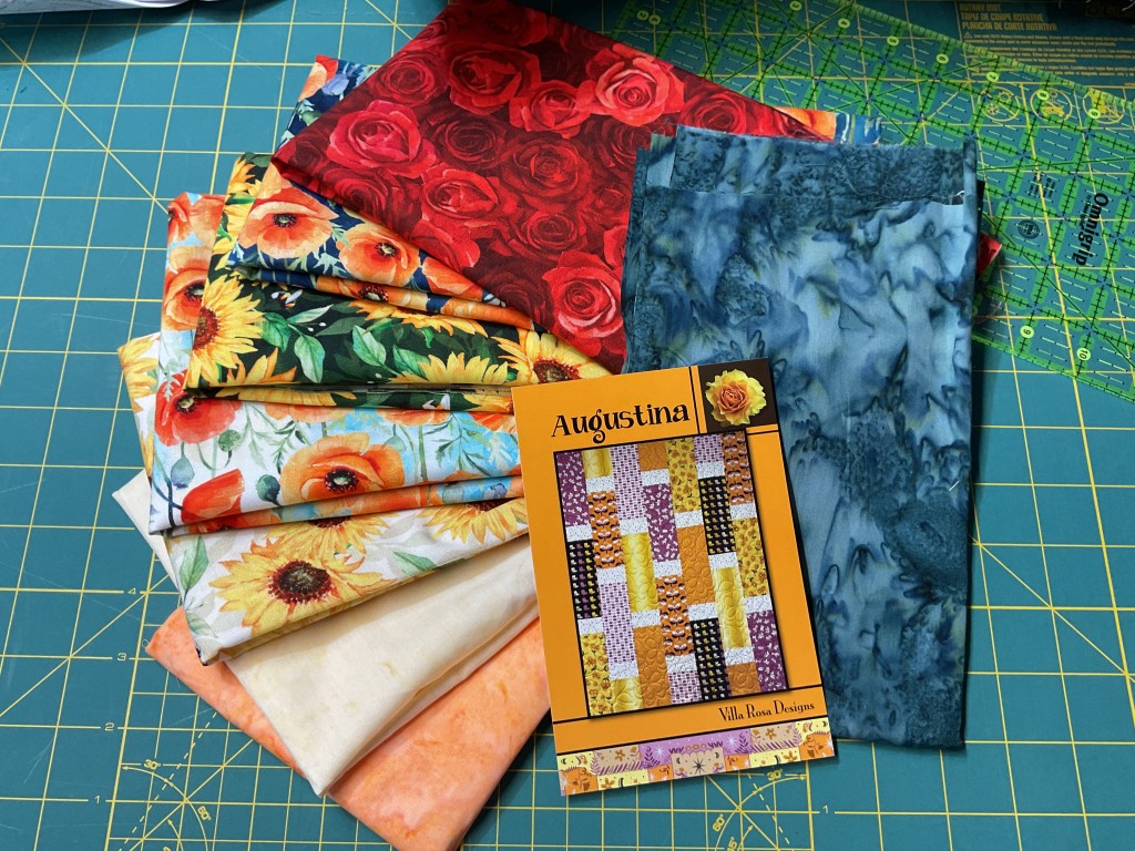 A collection of fabric pieces featuring floral patterns, including sunflowers and roses, stacked with an Augustina quilt pattern booklet on a cutting mat.