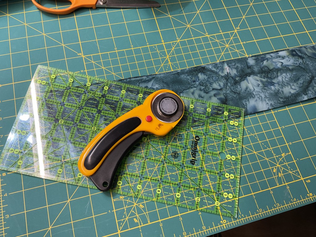 A rotary cutter, cutting mat, and ruler placed on a green cutting mat with yellow grid lines, with a strip of fabric in the background.