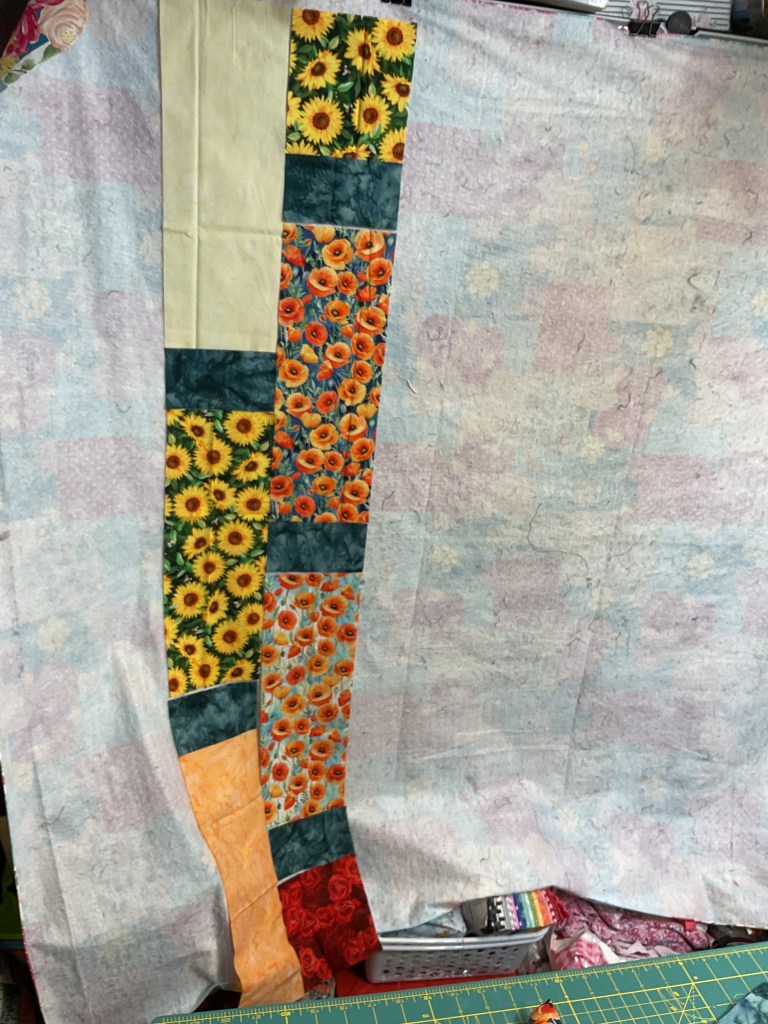 A colorful quilt top featuring sunflower and poppy floral patterns, laid out for sewing with a light blue background and various fabric strips.
