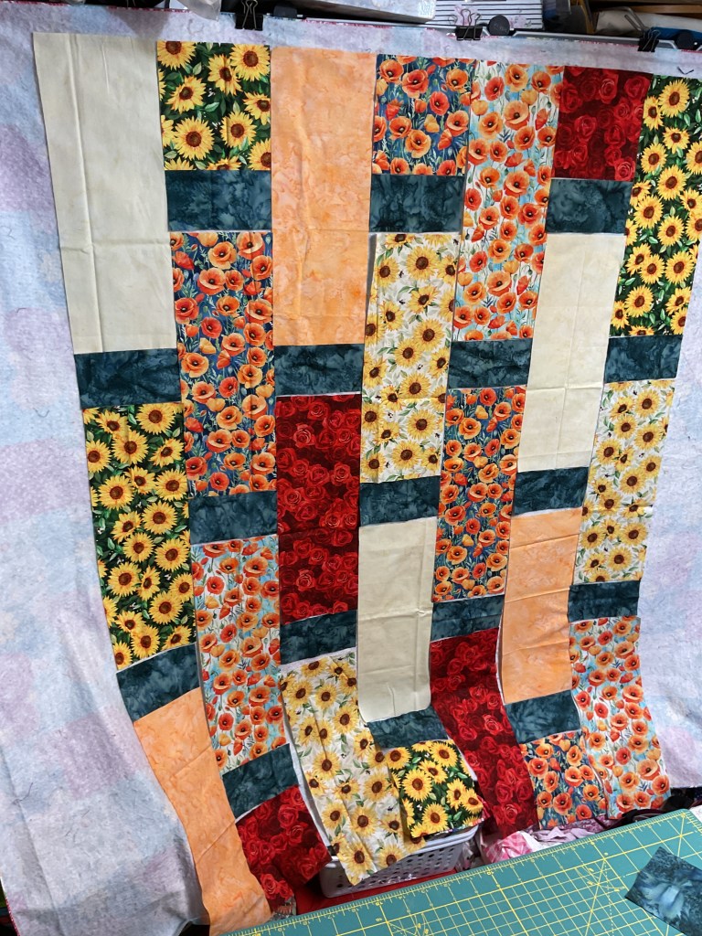 A layout of quilt fabric pieces showcasing bright floral patterns including sunflowers, poppies, and abstract designs, arranged in vertical strips with a green cutting mat below.