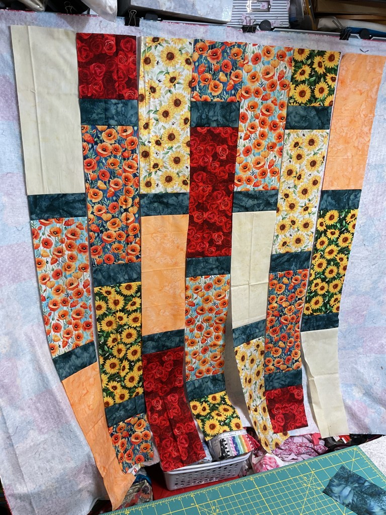 Vertical strips of colorful floral and solid fabrics are laid out for a quilt project, with a cutting mat visible below.