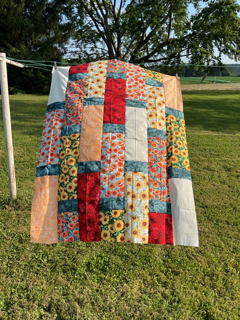 A colorful quilt featuring floral patterns and various fabrics, hanging on a clothesline outdoors with green grass and trees in the background.