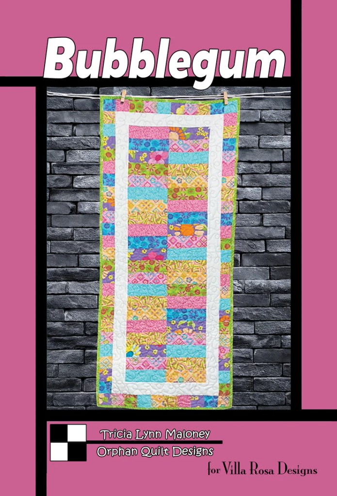 A colorful quilt named 'Bubblegum' designed by Tricia Lynn Maloney, featuring a patchwork layout with vibrant patterns, displayed against a textured black brick background.
