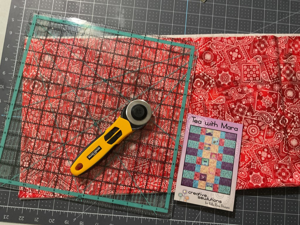 A rotary cutter and quilting ruler positioned on a cutting mat, with a vibrant red bandana print fabric and a quilt pattern titled 'Tea with Mara' by Creative Sewlutions.