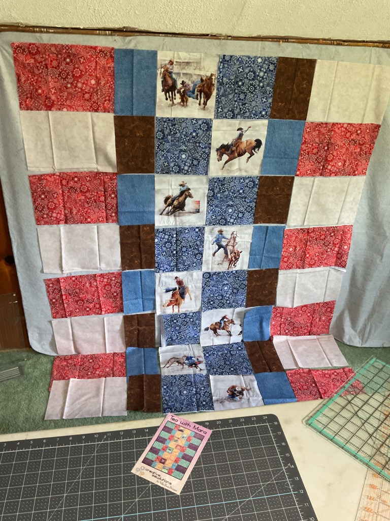 A colorful quilt layout featuring horse and rodeo-themed fabric squares, with red, blue, white, and brown patterned blocks arranged in a grid pattern on a design wall. A cutting mat and quilting ruler are visible in the foreground, along with a pattern card labeled 'Tea With Mara.'