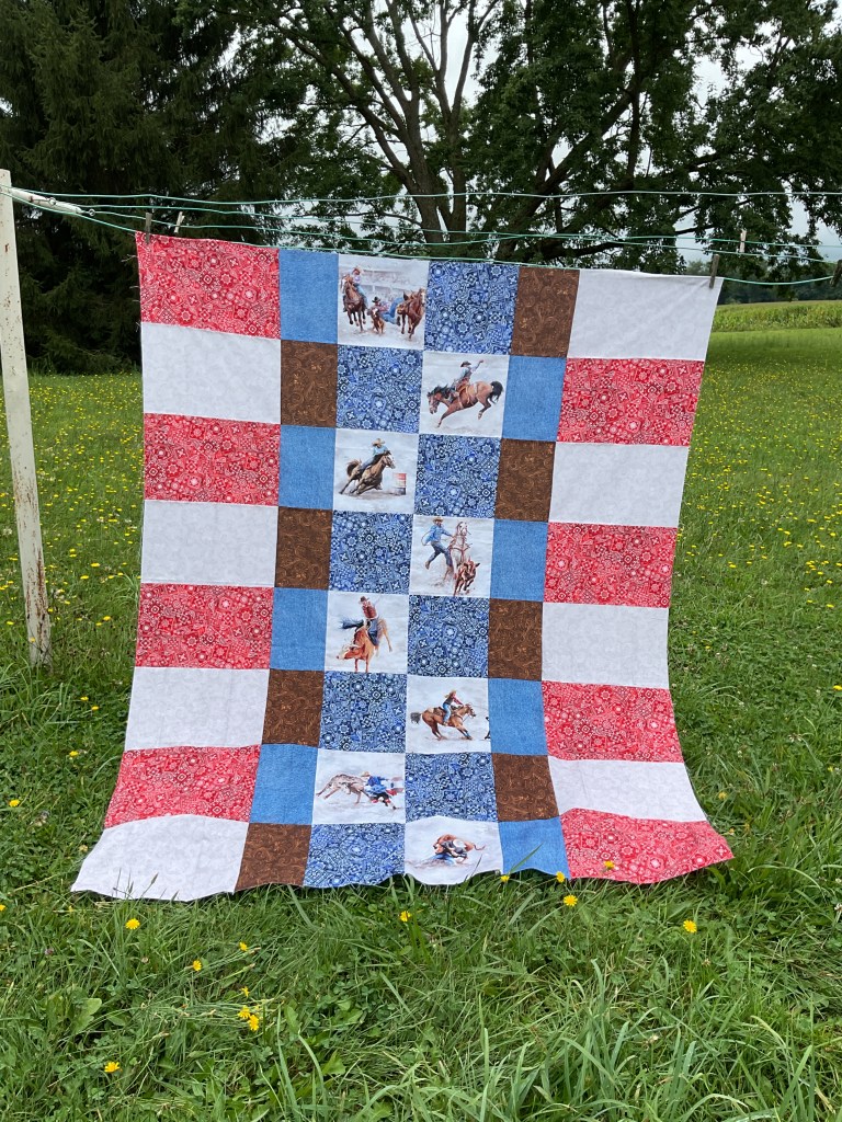 A colorful quilt with red, blue, brown, and white squares, featuring rodeo-themed prints, hanging on a clothesline in a grassy field.