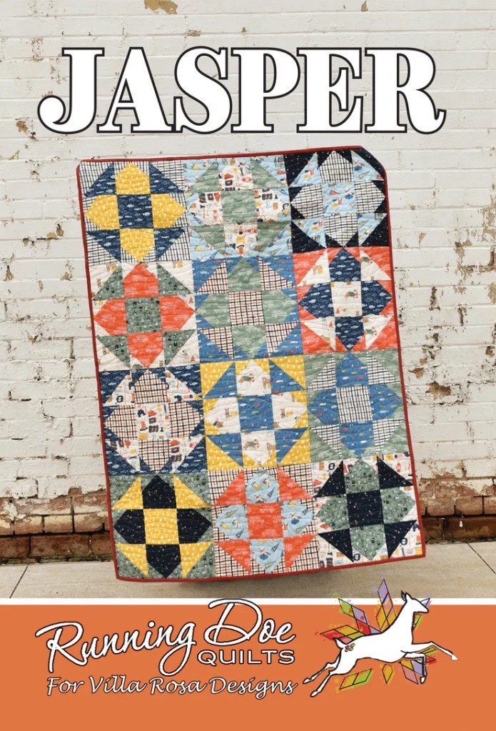 A colorful quilt named 'Jasper' featuring a variety of patterns in blue, green, yellow, and red fabric blocks, displayed against a brick wall background.