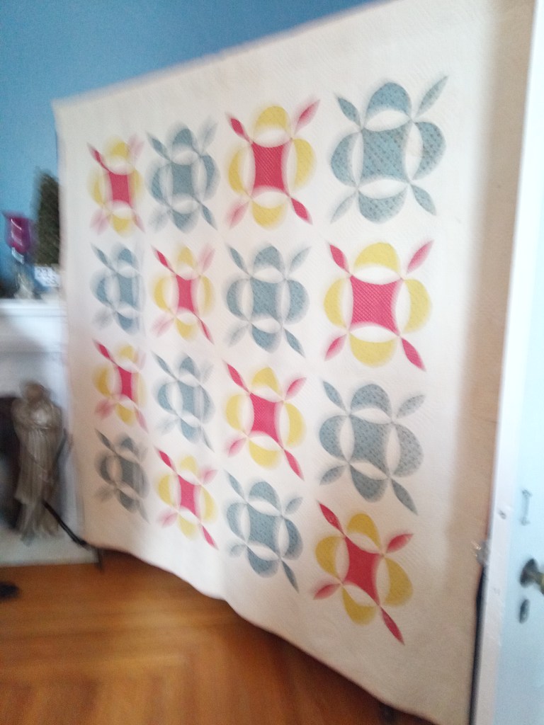 A colorful quilt featuring a pattern of interlocking shapes in pink, yellow, and blue, displayed against a softly colored wall.