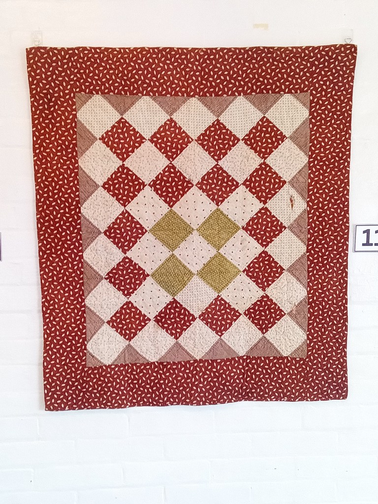 A quilt displayed on a white wall featuring a pattern of red, cream, and green squares with a stitched design.