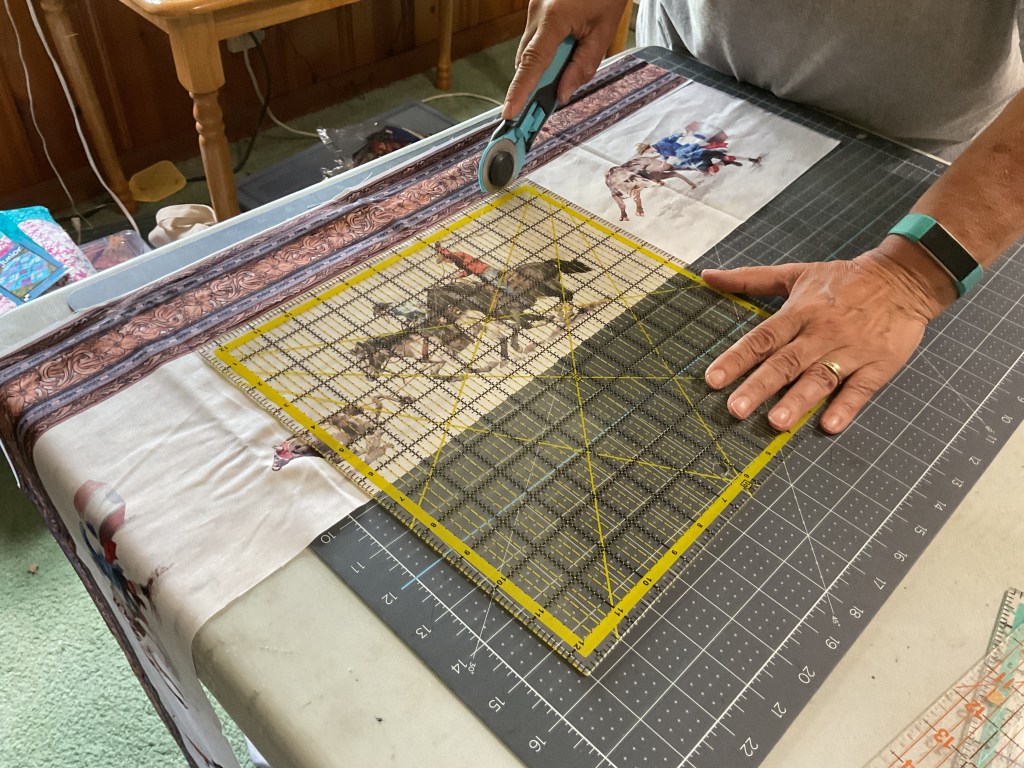 A person cutting fabric on a cutting mat with a rotary cutter, featuring printed rodeo scenes and decorative fabric borders.