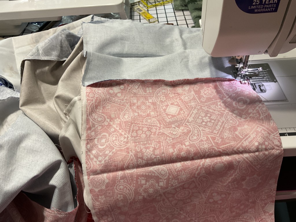 A sewing machine stitching together several pieces of fabric, including a patterned pink fabric and solid blue fabric, arranged on a sewing table.