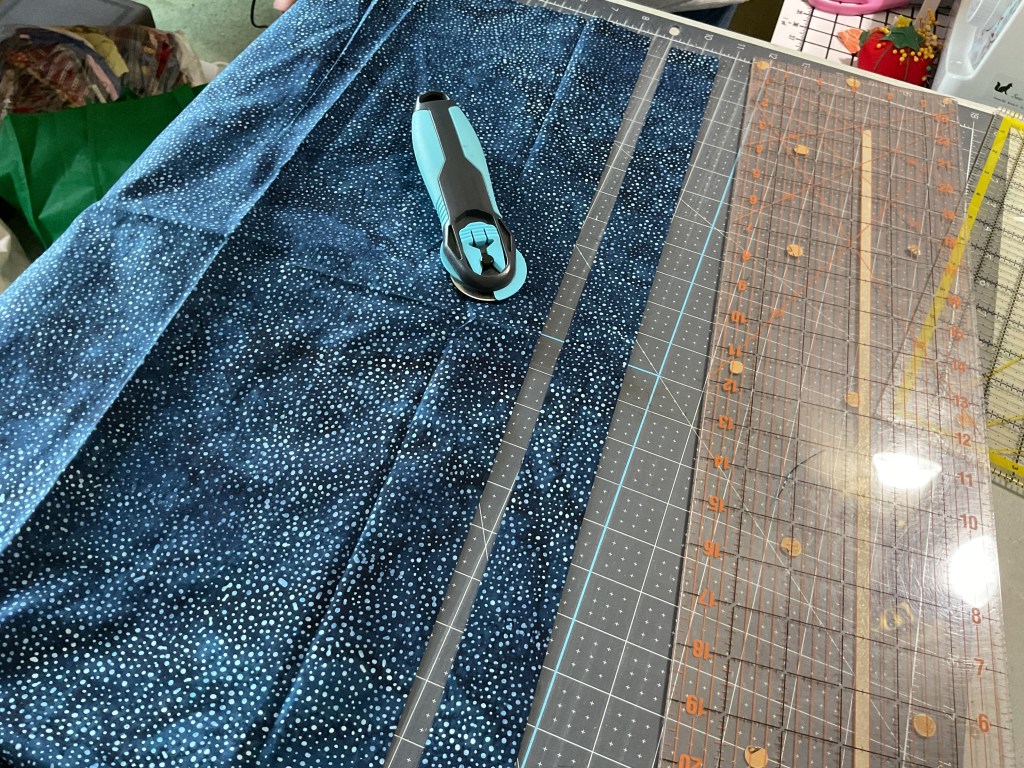 A rotary cutter is placed on a cutting mat with strips of navy blue fabric featuring white dot patterns, alongside a clear acrylic ruler for measuring.