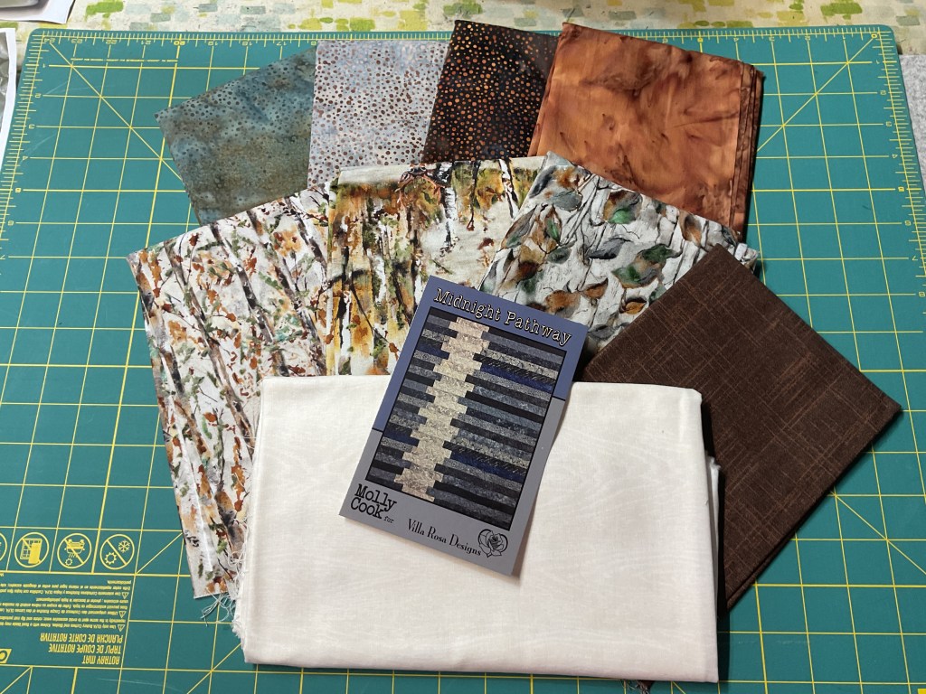 A collection of quilting fabrics laid out on a green cutting mat alongside a pattern card for the 'Midnight Pathway' quilt. The fabrics include various colors and patterns, while the card is clearly labeled with the quilt design details.