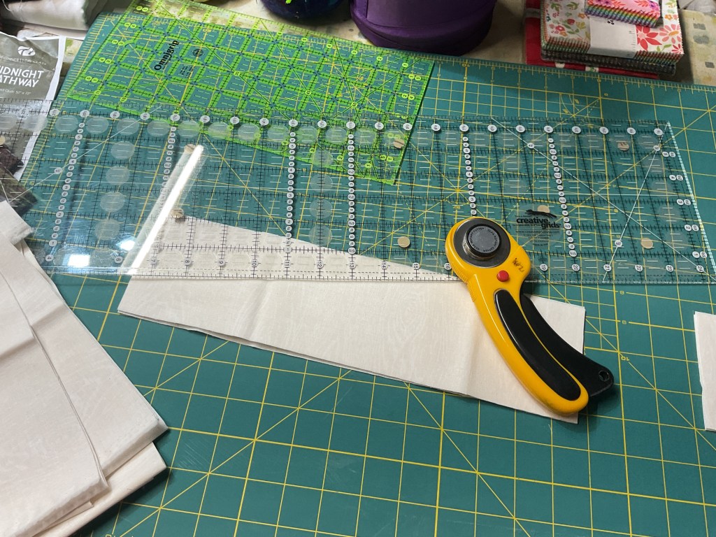 A quilting workspace featuring a rotary cutter, a clear acrylic ruler, and cut fabric pieces laid out on a green cutting mat.