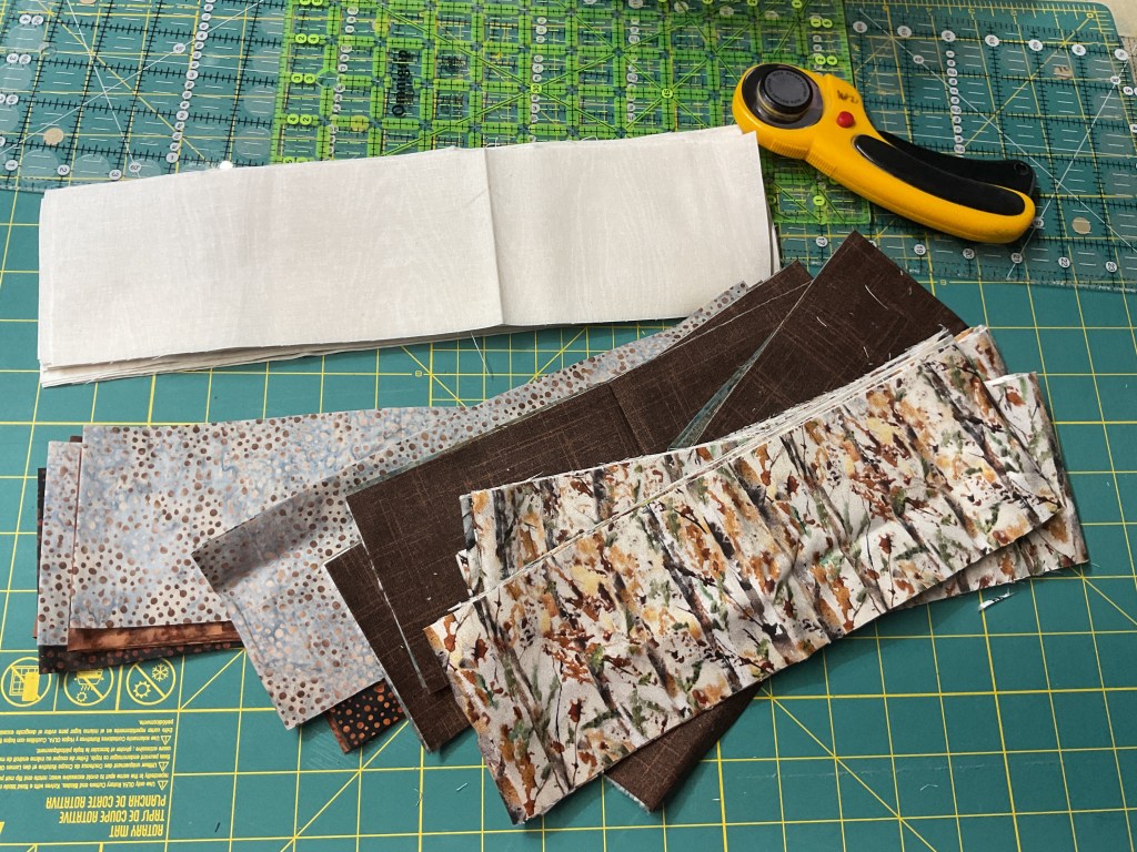 A collection of fabric strips in various patterns and colors, including light cream, earthy browns, and floral designs, arranged on a cutting mat alongside a rotary cutter and a ruler.