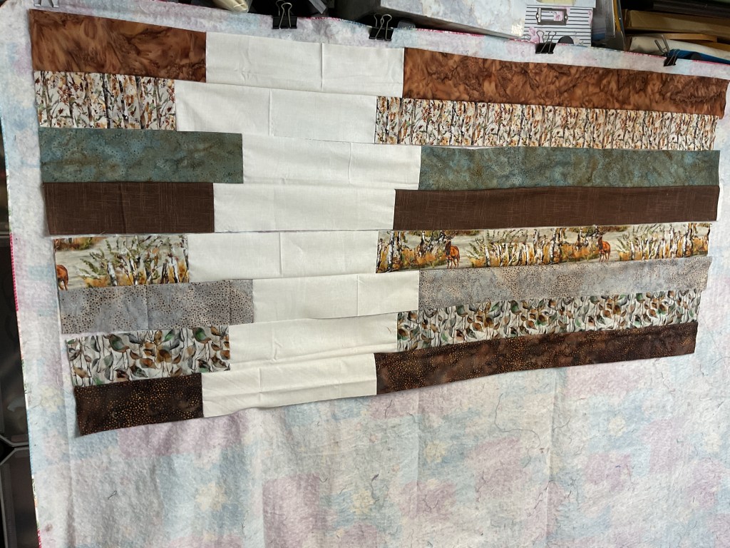 A partially completed quilt top featuring a variety of brown and blue fabric strips arranged in a modern design, displayed against a light-colored background.