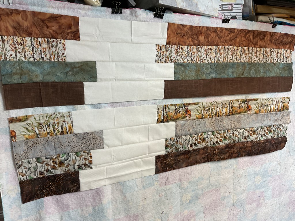 A patchwork quilt top featuring a variety of earthy fabric strips arranged in horizontal rows, showcasing a mix of patterns and colors, including browns, creams, and soft greens.