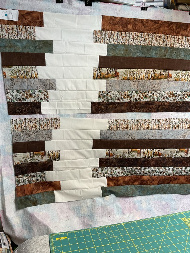 A partially completed quilt top displayed on a design wall, featuring alternating strips of earthy fabrics in various shades of brown, cream, and teal, alongside printed fabric depicting a nature scene with deer.