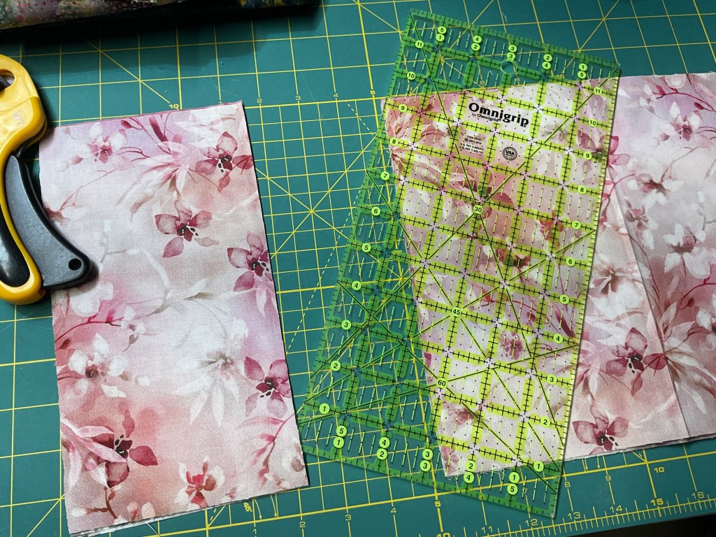 A cutting mat with a rotary cutter and floral fabric pieces for quilting. A ruler is positioned on top of the fabric, indicating measurement marks.