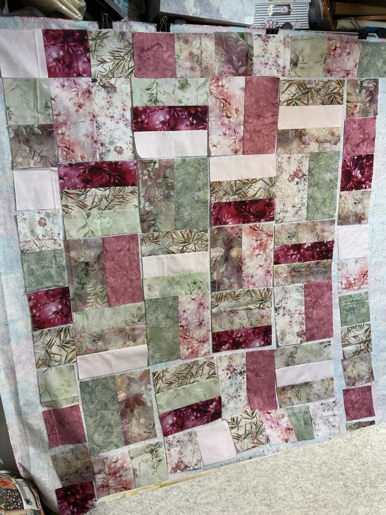 A quilt top layout featuring various floral and patterned fabric pieces in shades of green, pink, and mauve, arranged in a patchwork design.