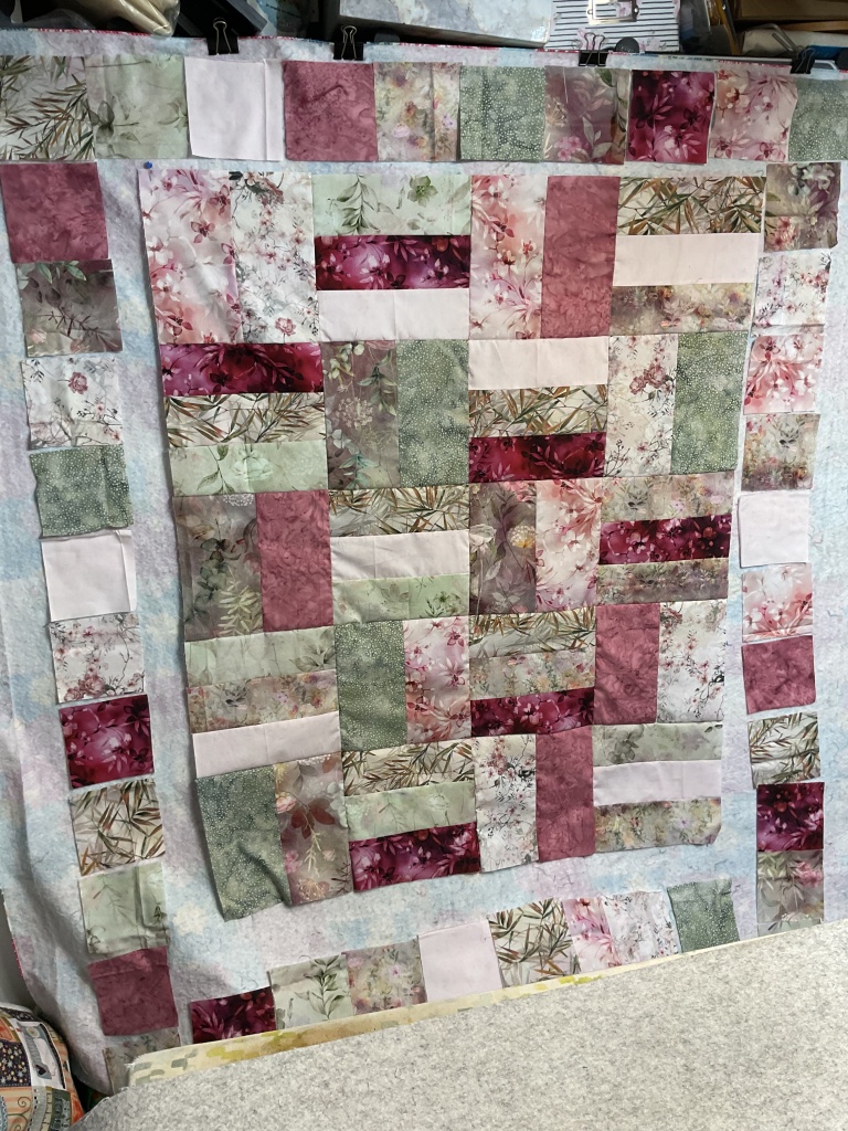 A partially completed quilt top featuring a variety of floral and patterned fabrics in shades of pink, green, and white, arranged in a block design on a light background.
